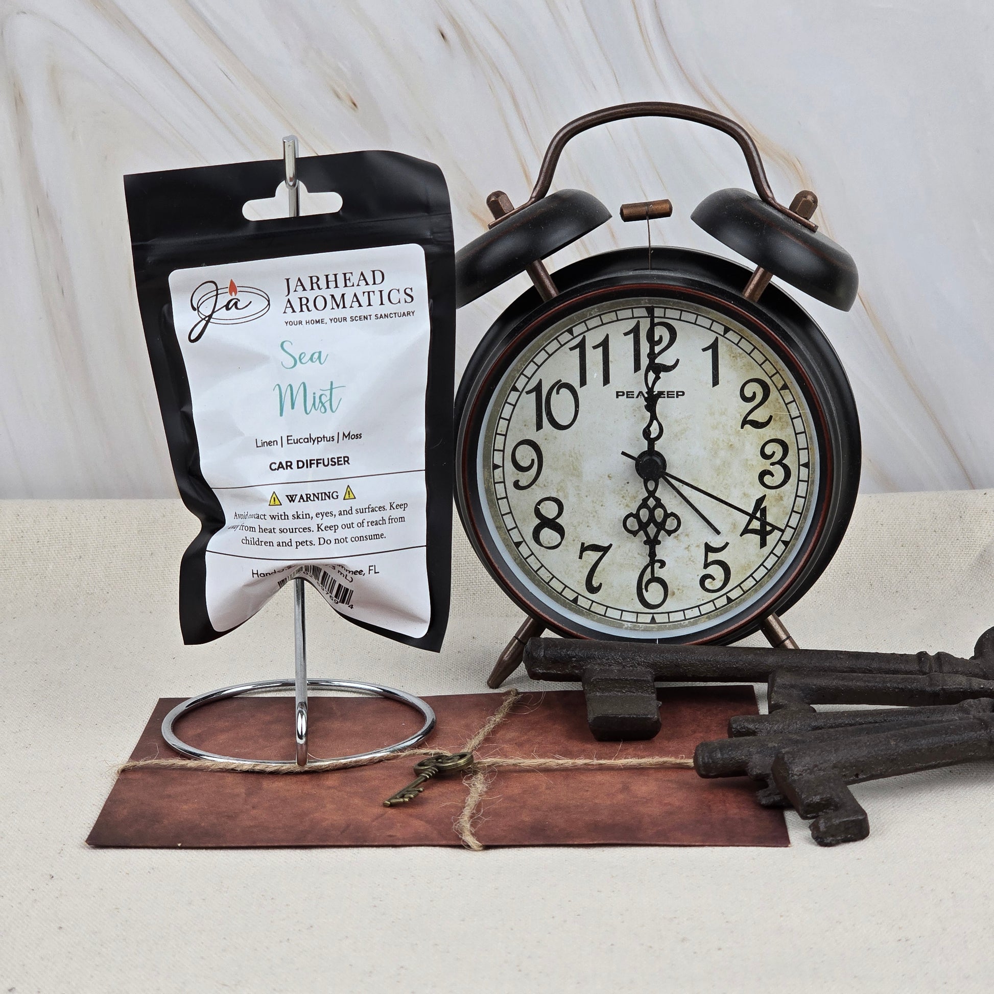 A packet of Jarhead Aromatics Sea Mist Car Diffuser | Clean Coastal & Spa-Inspired Car Air Freshener hangs on a metal stand beside a vintage black alarm clock, old keys, and a brown envelope on a neutral surface.