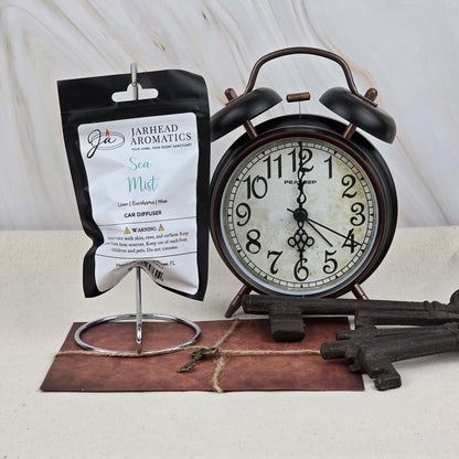 A packet of Jarhead Aromatics Sea Mist Car Diffuser | Clean Coastal & Spa-Inspired Car Air Freshener hangs on a metal stand beside a vintage black alarm clock, old keys, and a brown envelope on a neutral surface.