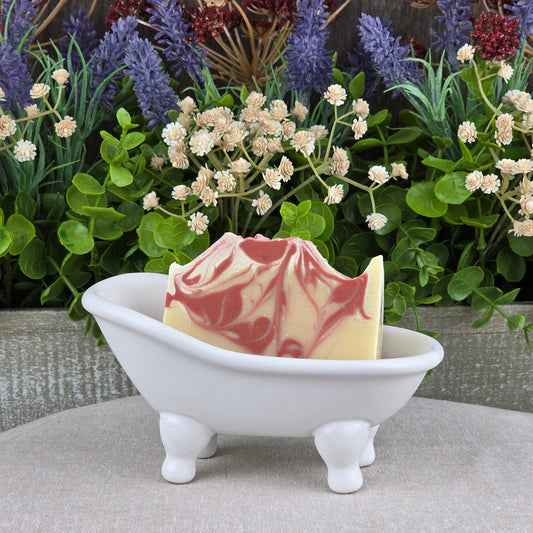 The Sea Moss + Aloe Handmade Artisan Soap by Jarhead Aromatics, featuring red and white swirls, rests in a small white clawfoot bathtub dish with lush greenery and blooming flowers in the background.