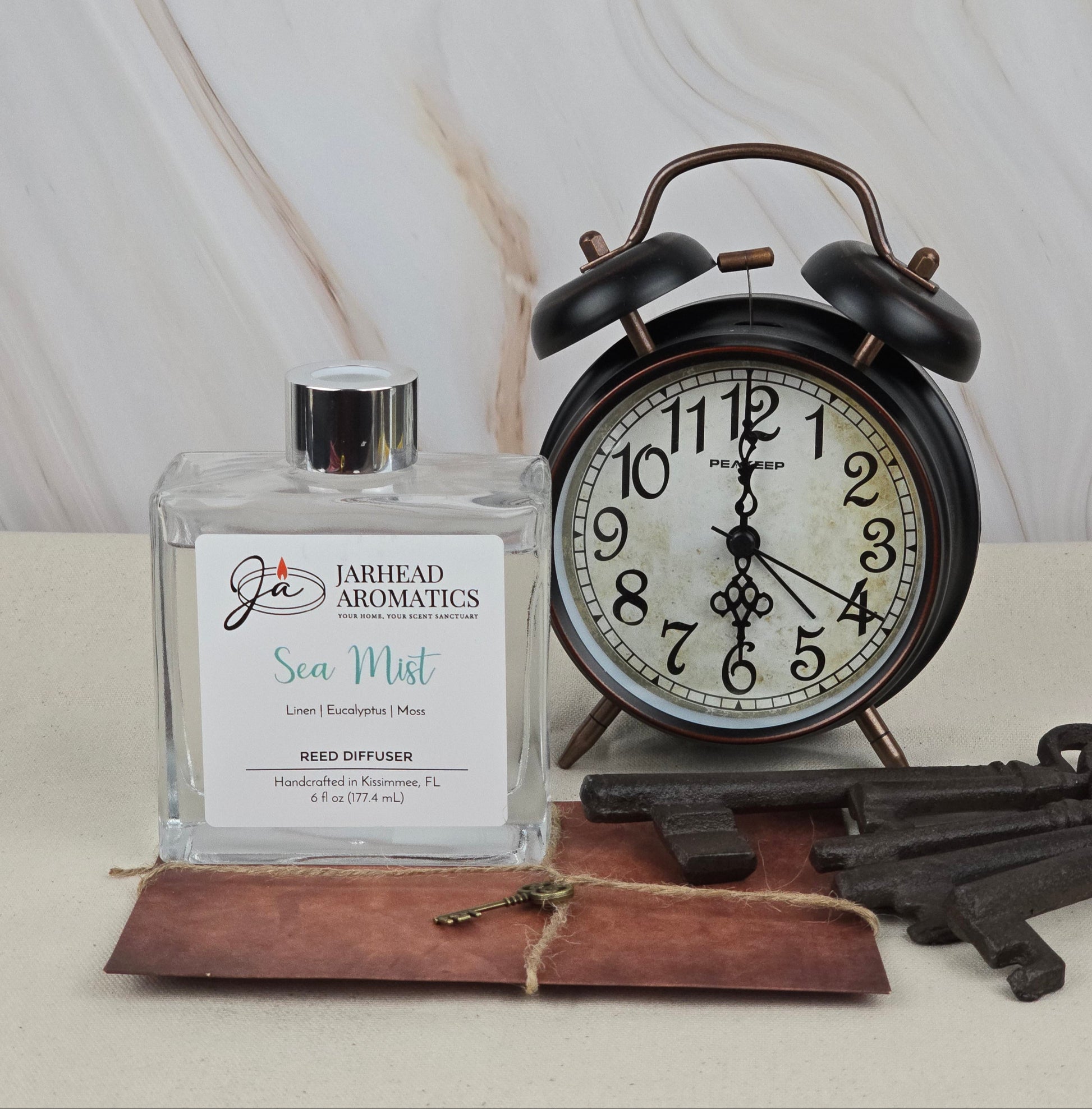 A square Jarhead Aromatics Sea Mist reed diffuser with a white label featuring blue and red text sits next to a vintage black alarm clock. Nearby, old-fashioned keys rest on a small leather notebook, adding charm to the peaceful scene.