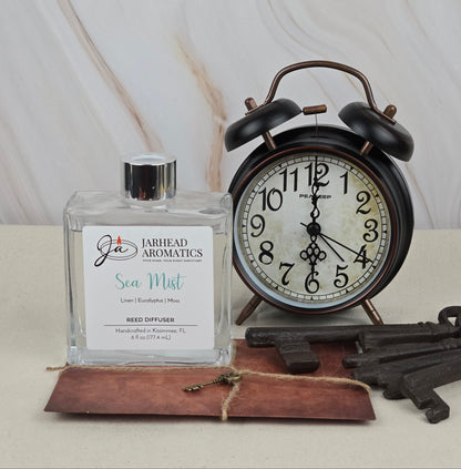 A square Jarhead Aromatics Sea Mist reed diffuser with a white label featuring blue and red text sits next to a vintage black alarm clock. Nearby, old-fashioned keys rest on a small leather notebook, adding charm to the peaceful scene.