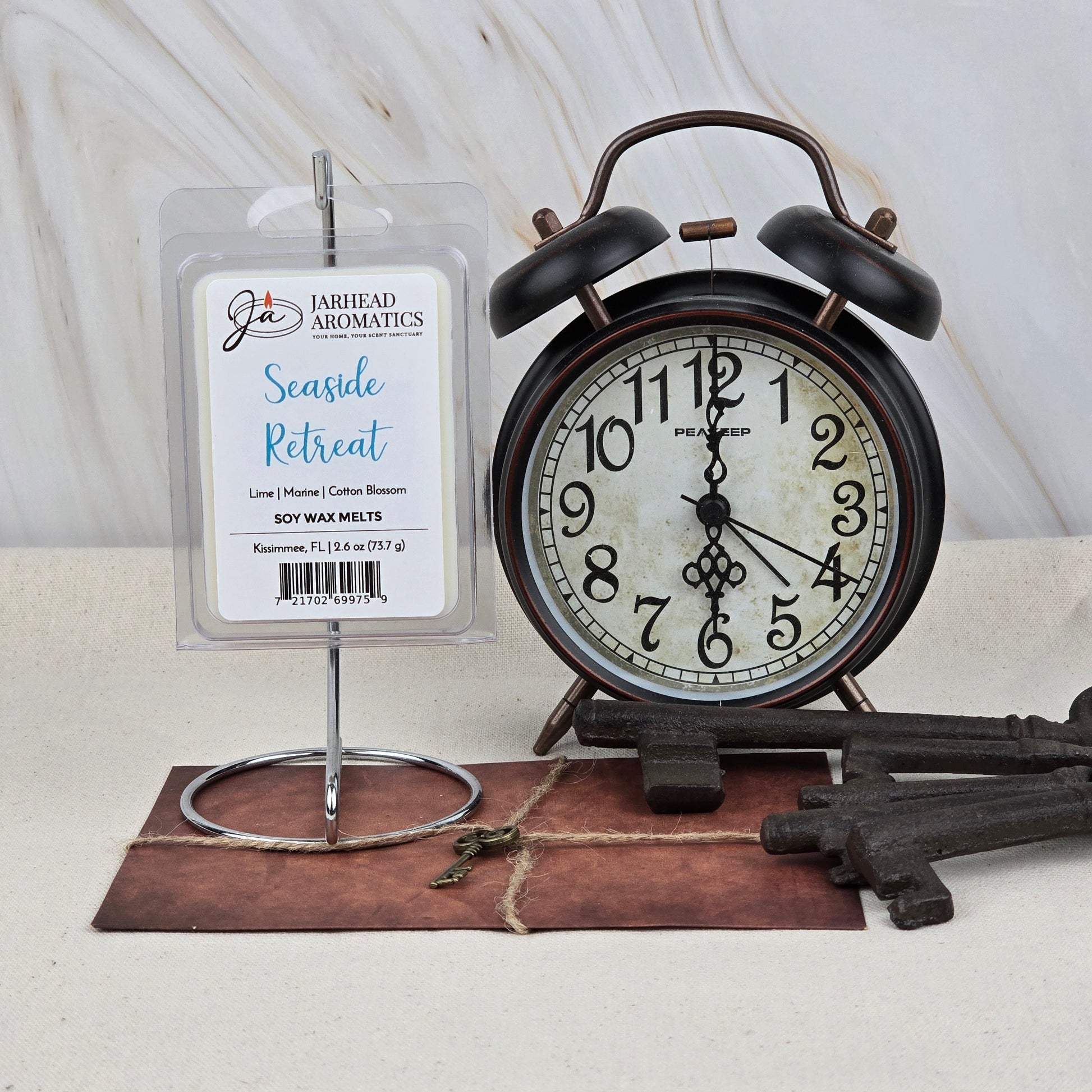 A package of Jarhead Aromatics Seaside Retreat Soy Wax Melt | Fresh Coastal Citrus & Ocean Breeze sits on a metal holder beside a vintage black alarm clock and keys atop a brown folder, against a light marble background, offering long-lasting fragrance.