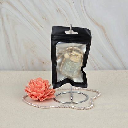A black ziplock pouch from Jarhead Aromatics sits on a metal holder, showcasing the Island Hibiscus Car Diffuser | Tropical Floral & Melon inside. In front are a pink artificial flower and pearls on a light beige surface, set against marble.