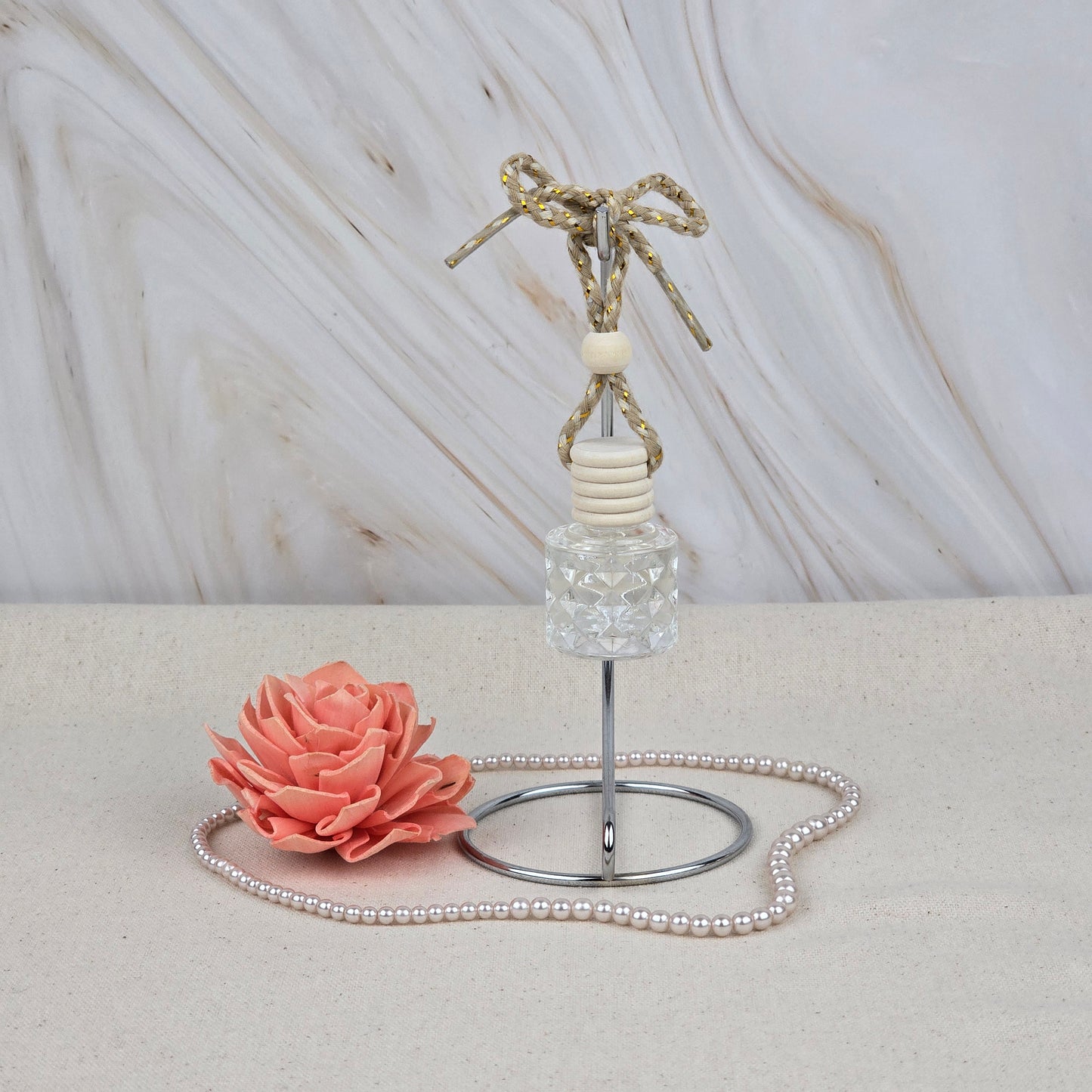 A Jarhead Aromatics Island Hibiscus Car Diffuser | Tropical Floral & Melon Car Air Freshener with a rope rests on a metal stand next to a pink decorative flower and pearls on a light beige surface, bringing a tropical scent.