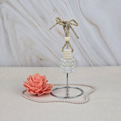A Jarhead Aromatics Island Hibiscus Car Diffuser | Tropical Floral & Melon Car Air Freshener with a rope rests on a metal stand next to a pink decorative flower and pearls on a light beige surface, bringing a tropical scent.
