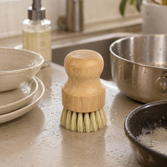 A Jarhead Aromatics Bamboo Handle Sisal Fiber Palm Scrubber Brush | Natural Kitchen Dish & Produce Cleaning Brush stands on a wet kitchen countertop beside stacked plates, a sudsy pan, a metal bowl, and a soap dispenser—an eco-friendly cleaning tool ideal for effortless dishwashing.