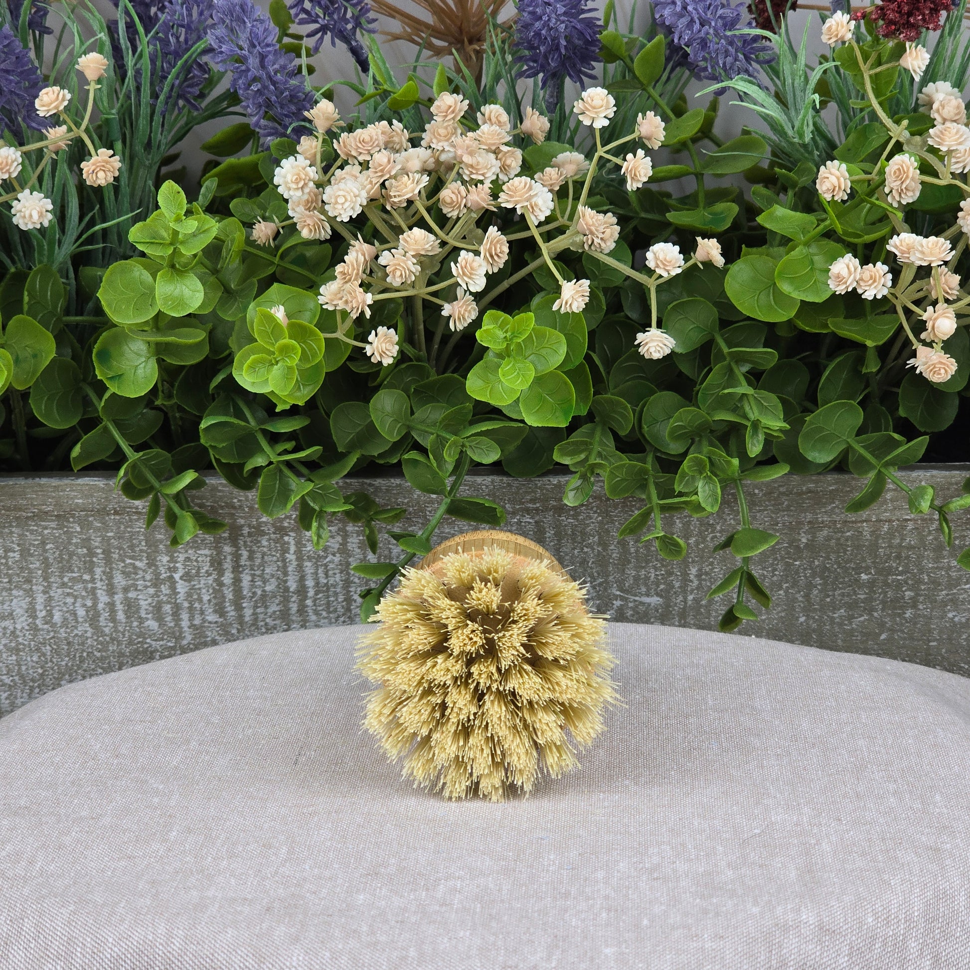 The Jarhead Aromatics Bamboo Handle Sisal Fiber Palm Scrubber Brush, a natural kitchen dish and produce cleaning brush, is displayed on a beige fabric with green leaves and white and purple flowers in the background.