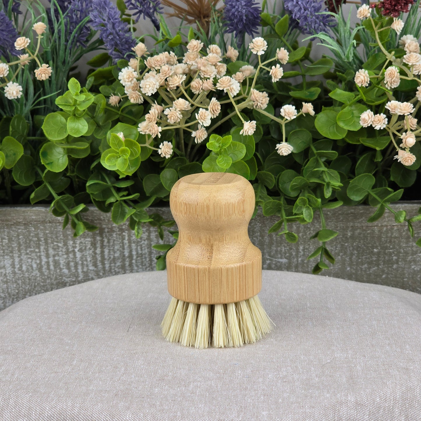 The Jarhead Aromatics Bamboo Handle Sisal Fiber Palm Scrubber Brush stands upright on gray fabric, surrounded by greenery and white flowers—a natural and eco-friendly kitchen dish and produce cleaning brush.