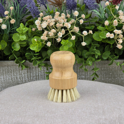 The Jarhead Aromatics Bamboo Handle Sisal Fiber Palm Scrubber Brush stands upright on gray fabric, surrounded by greenery and white flowers—a natural and eco-friendly kitchen dish and produce cleaning brush.