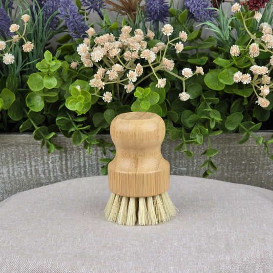 The Jarhead Aromatics Bamboo Handle Sisal Fiber Palm Scrubber Brush stands upright on gray fabric, surrounded by greenery and white flowers—a natural and eco-friendly kitchen dish and produce cleaning brush.