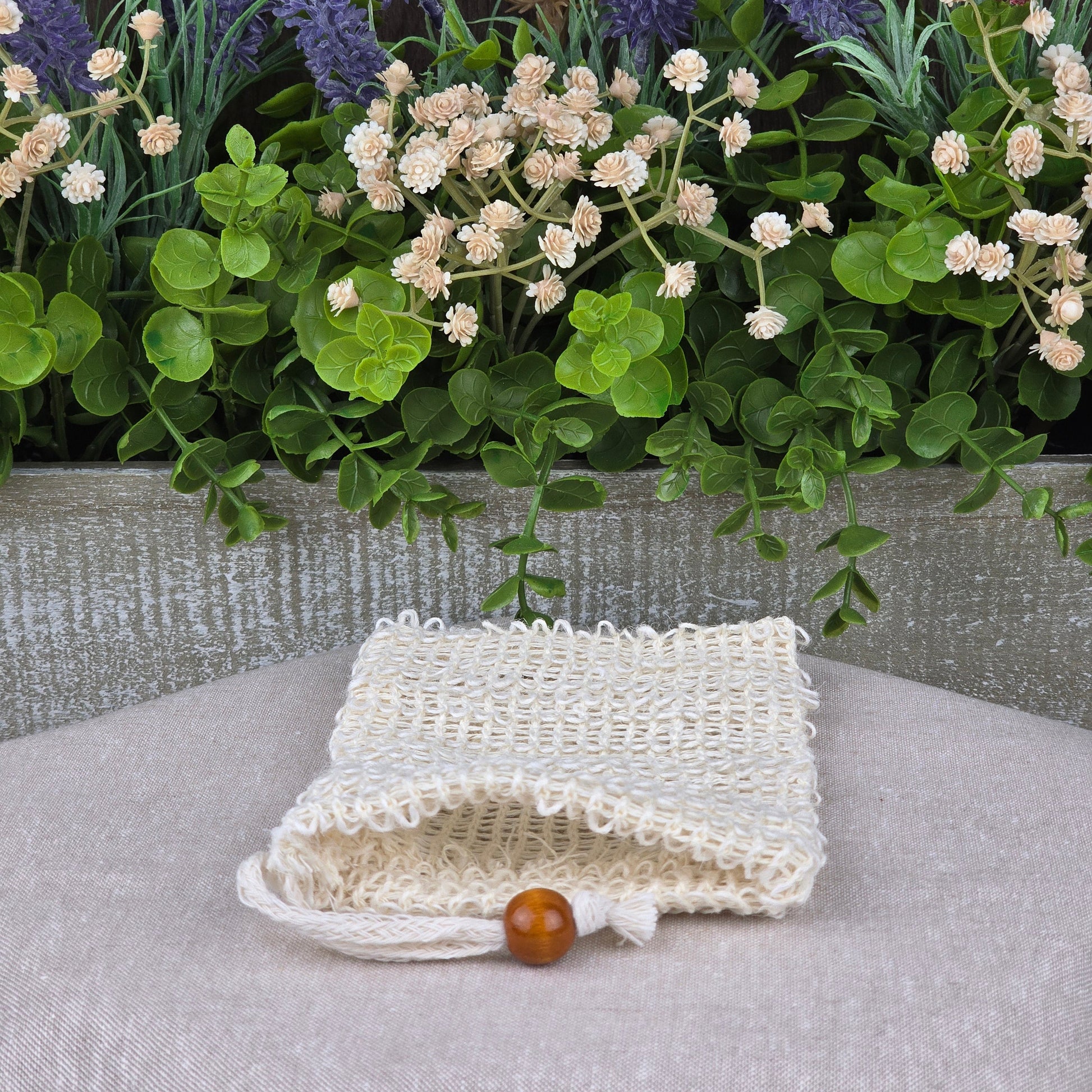 The Sisal Fiber Natural Soap Saver Bag by Jarhead Aromatics, featuring a wooden bead drawstring, rests on a beige surface with lush green leaves and purple, white, and burgundy artificial flowers against a wooden background.