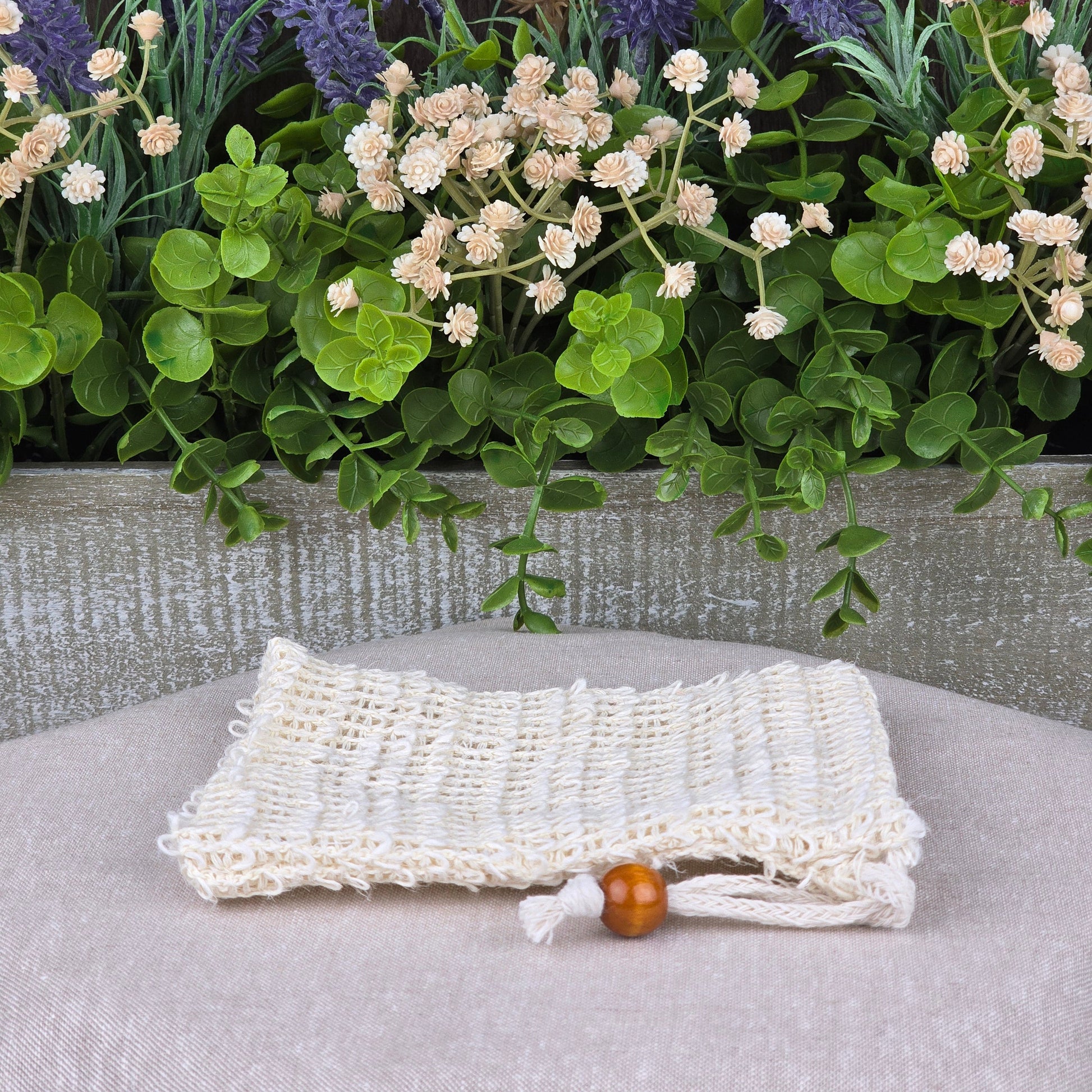 The Sisal Fiber Natural Soap Saver Bag by Jarhead Aromatics, a cream-colored exfoliating pouch with a wooden bead, rests on beige beside green foliage and purple and red flowers, set against a gray wood backdrop.