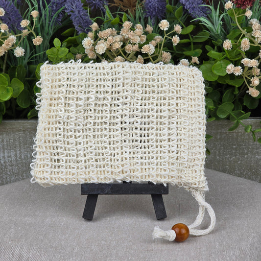 The Jarhead Aromatics Sisal Fiber Natural Soap Saver Bag, an exfoliating soap pouch and zero-waste soap holder, is displayed on a black easel with artificial flowers and greenery in the background for a natural vibe.