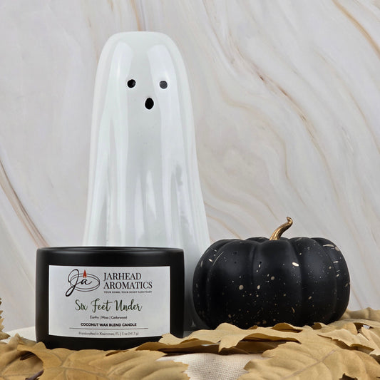 A white ceramic ghost, a black pumpkin, and Jarhead Aromatics’ Six Feet Under - Coconut Wax Blend Candle rest on beige fabric with decaying leaves, set against a marble background.