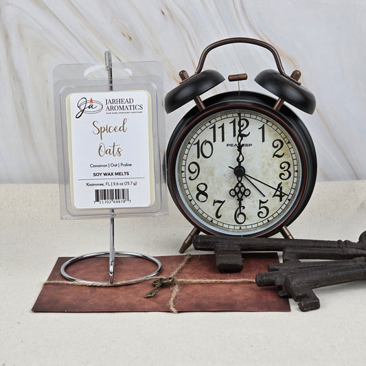 Jarhead Aromatics Spiced Oats Soy Wax Melt, with a warm oatmeal brown sugar scent, sits on a metal holder beside a vintage black alarm clock and large keys atop a brown envelope on a light marble background.