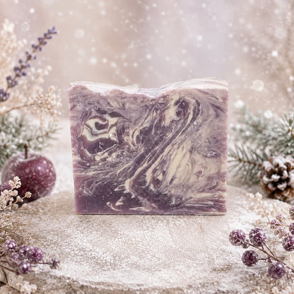 A bar of Jarhead Aromatics Sugar Plum Handmade Coconut Milk Artisan Soap with purple and cream swirls sits on a frosted stone surface surrounded by pine branches, pinecones, and berries.