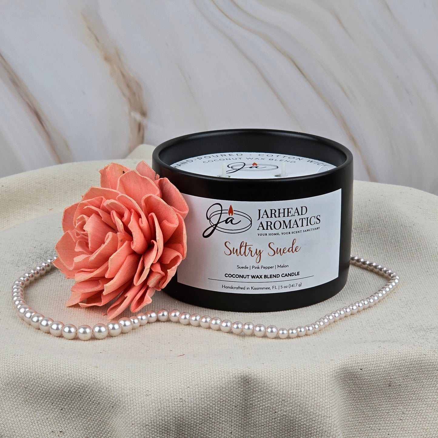 A Jarhead Aromatics Sultry Suede Coconut Wax Blend Candle, scented with suede, tobacco, and fruit, rests in a sleek black tin beside a coral-hued wooden flower and pearl necklace on neutral textured fabric.