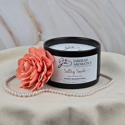 A Jarhead Aromatics Sultry Suede Coconut Wax Blend Candle, scented with suede, tobacco, and fruit, rests in a sleek black tin beside a coral-hued wooden flower and pearl necklace on neutral textured fabric.