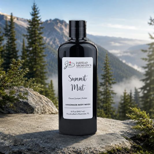 A black bottle of Jarhead Aromatics Summit Mist Handmade Body Wash | Moisturizing Body Wash with Organic Aloe & Plant-Based Oils rests on a rock, set against a scenic backdrop of forest, mountains, and mist beneath a clear sky.
