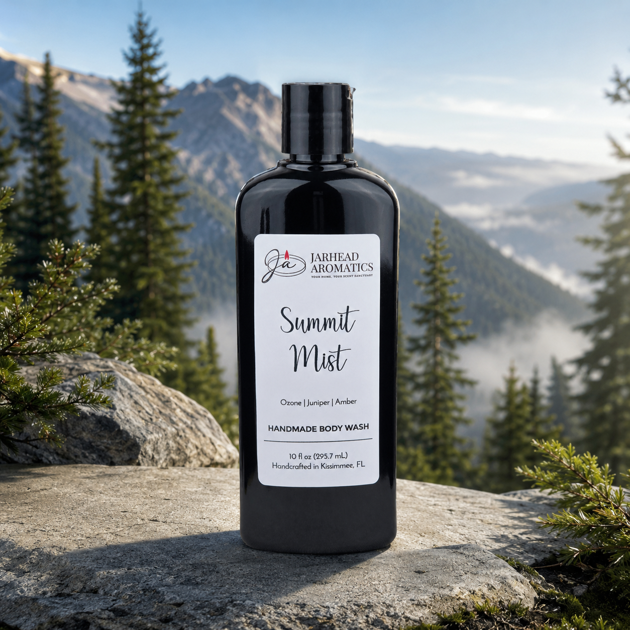 A black bottle of Jarhead Aromatics Summit Mist Handmade Body Wash with organic aloe and plant-based oils stands on a rock, set against misty pines and mountains. The label features notes of ozone, juniper, and amber.