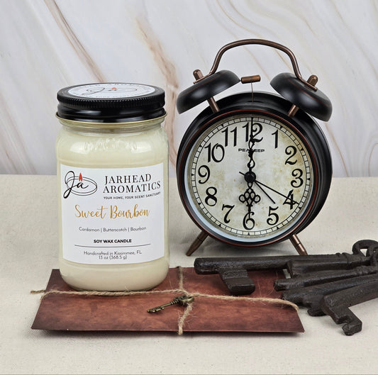 A Jarhead Aromatics Sweet Bourbon soy wax candle with rich cardamom, butterscotch, and bourbon scents sits next to a vintage alarm clock. Its warm home fragrance complements the nearby old keys and brown vintage paper on a neutral surface.