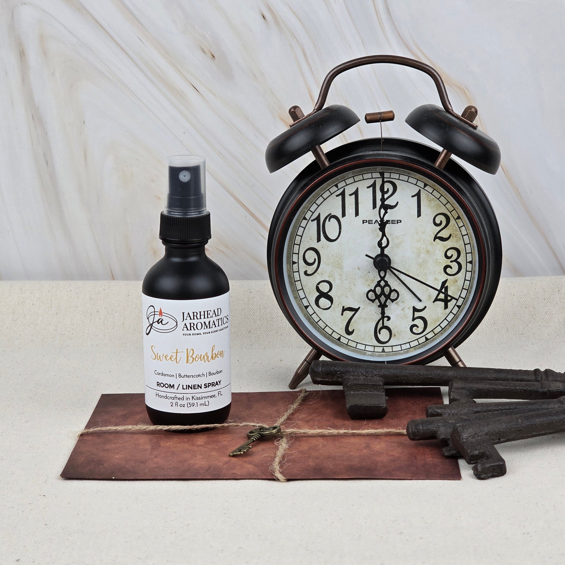 A bottle of Jarhead Aromatics Sweet Bourbon Room & Linen Spray, a home fragrance with bourbon, brown sugar, and oak, is placed beside a vintage black alarm clock, four antique-style keys, and a string-tied envelope on a neutral surface.