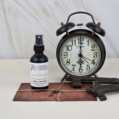 A bottle of Jarhead Aromatics Sweet Bourbon Room & Linen Spray, a home fragrance with bourbon, brown sugar, and oak, is placed beside a vintage black alarm clock, four antique-style keys, and a string-tied envelope on a neutral surface.