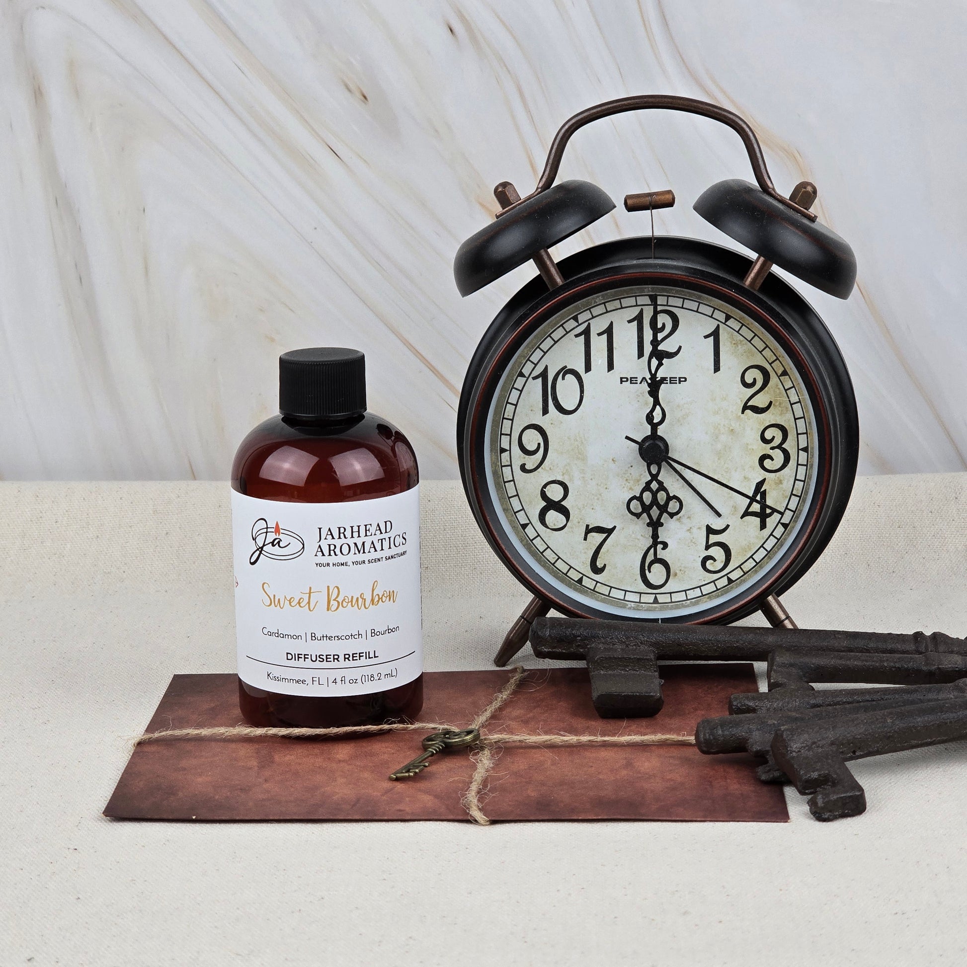 A vintage alarm clock, large decorative keys, and a small key rest on a leather-bound surface against light marble. The scene is infused with the inviting scent of Sweet Bourbon Diffuser Refill by Jarhead Aromatics—bourbon, brown sugar & oak.