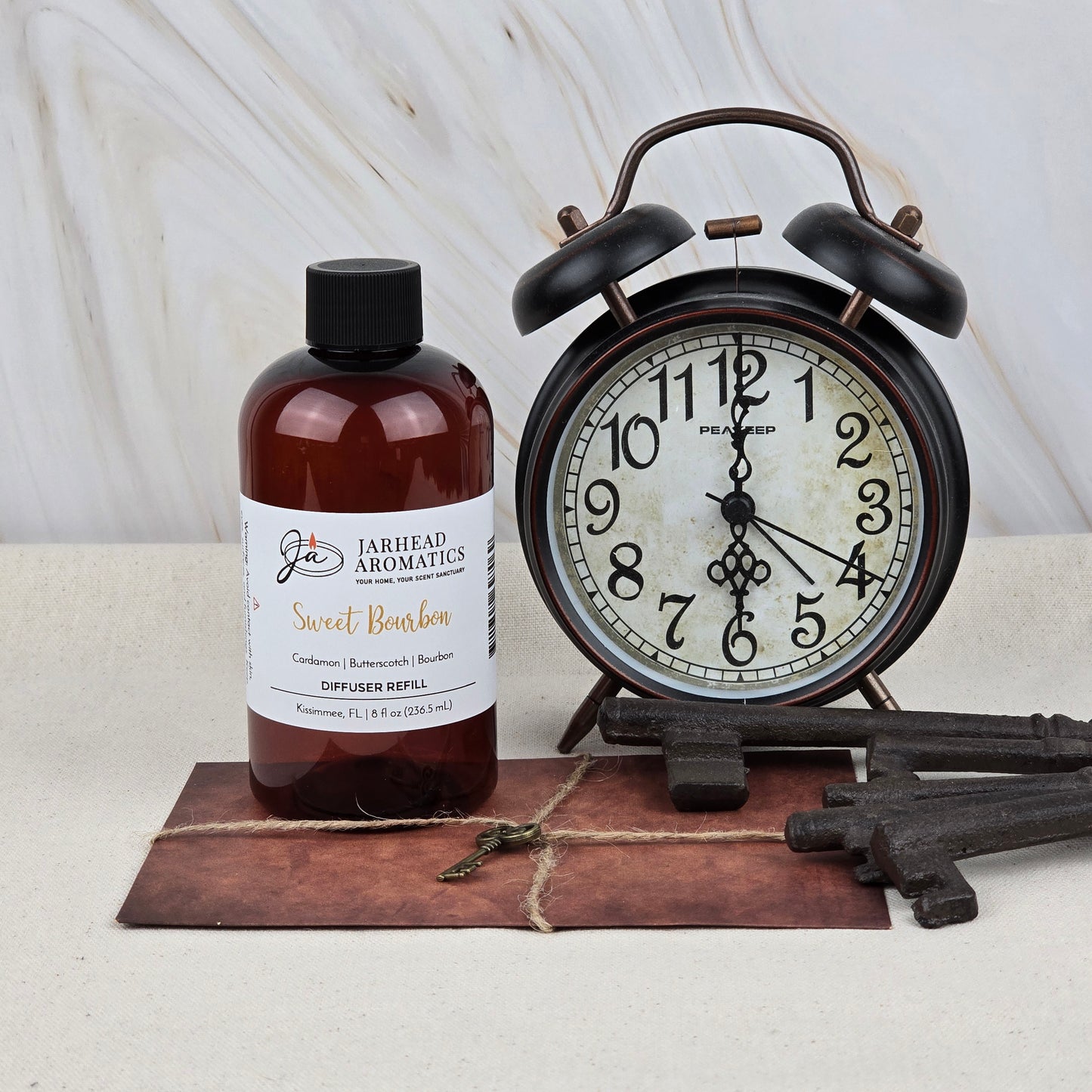 A bottle of Jarhead Aromatics Sweet Bourbon Diffuser Refill, a vintage black alarm clock, three old-fashioned keys, and a leather-bound notebook create a cozy scene—ideal for filling your home with bourbon, brown sugar & oak fragrance.