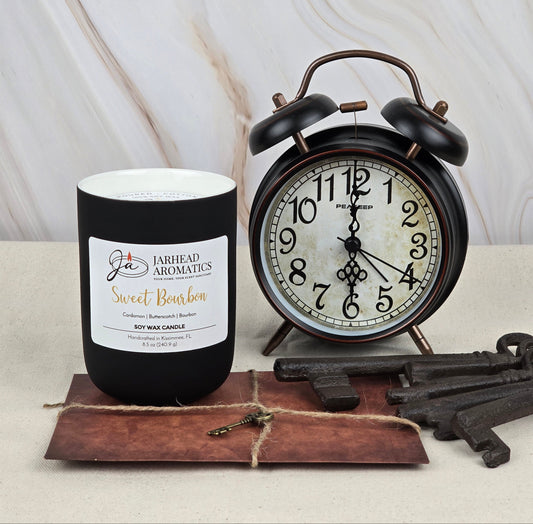 A Sweet Bourbon - Soy Wax Candle by Jarhead Aromatics emits a rich bourbon fragrance, elegantly set next to a classic black alarm clock on beige. Antique metal keys accompany them, and the brown vintage paper enhances the cozy home scent ambiance.