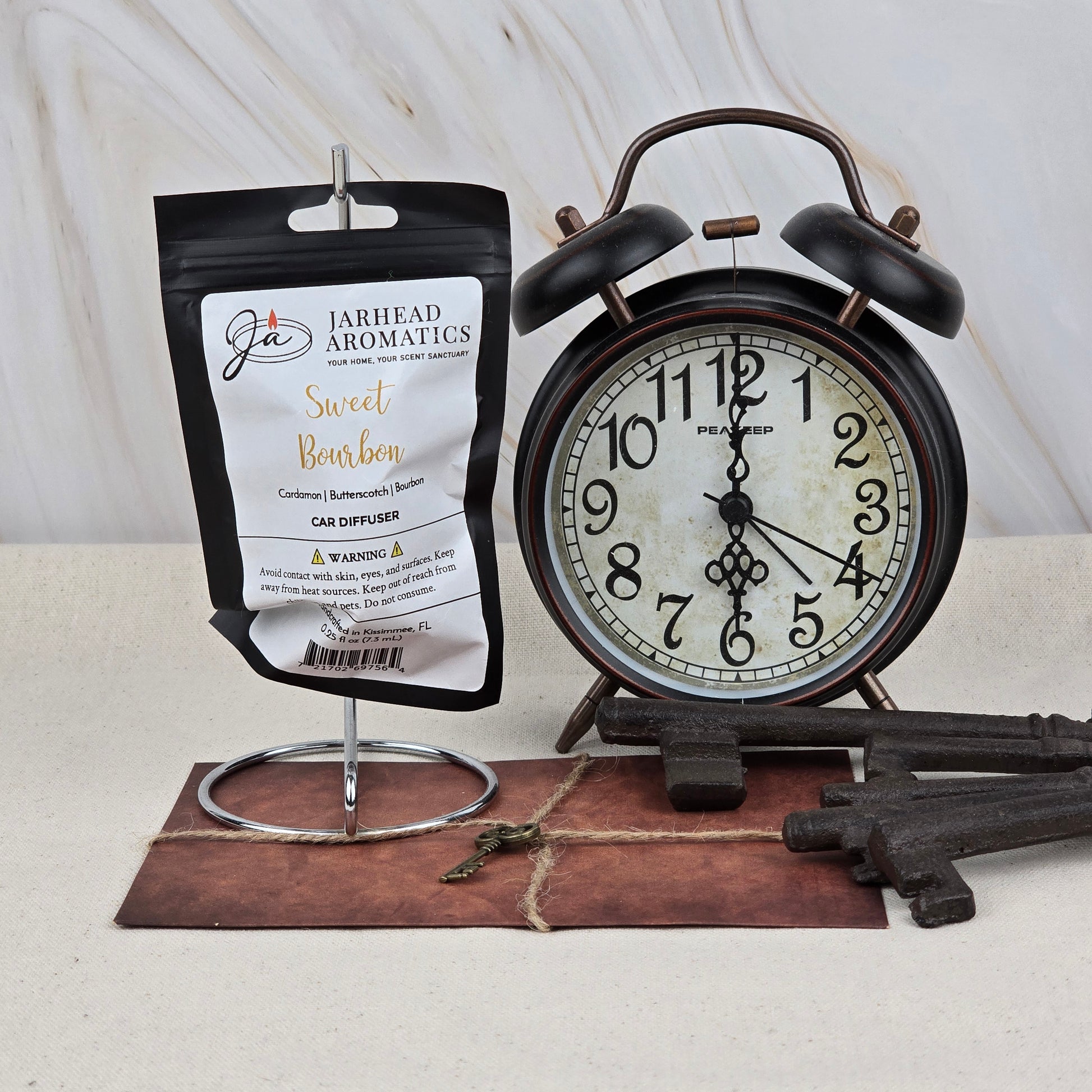 A Jarhead Aromatics Sweet Bourbon Car Diffuser, scented with bourbon, brown sugar, and oak, hangs on a stand beside a vintage black alarm clock, antique keys, and a brown notepad with a key on top against a neutral background.