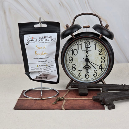 A Jarhead Aromatics Sweet Bourbon Car Diffuser, scented with bourbon, brown sugar, and oak, hangs on a stand beside a vintage black alarm clock, antique keys, and a brown notepad with a key on top against a neutral background.