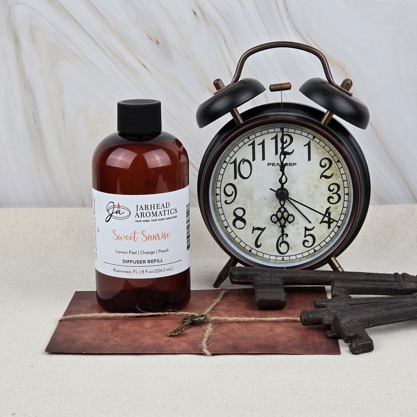 A brown alarm clock sits next to a bottle of Jarhead Aromatics Sweet Sunrise Diffuser Refill | Citrus, Agave & Peach Home Fragrance Oil on a rustic surface with vintage keys.