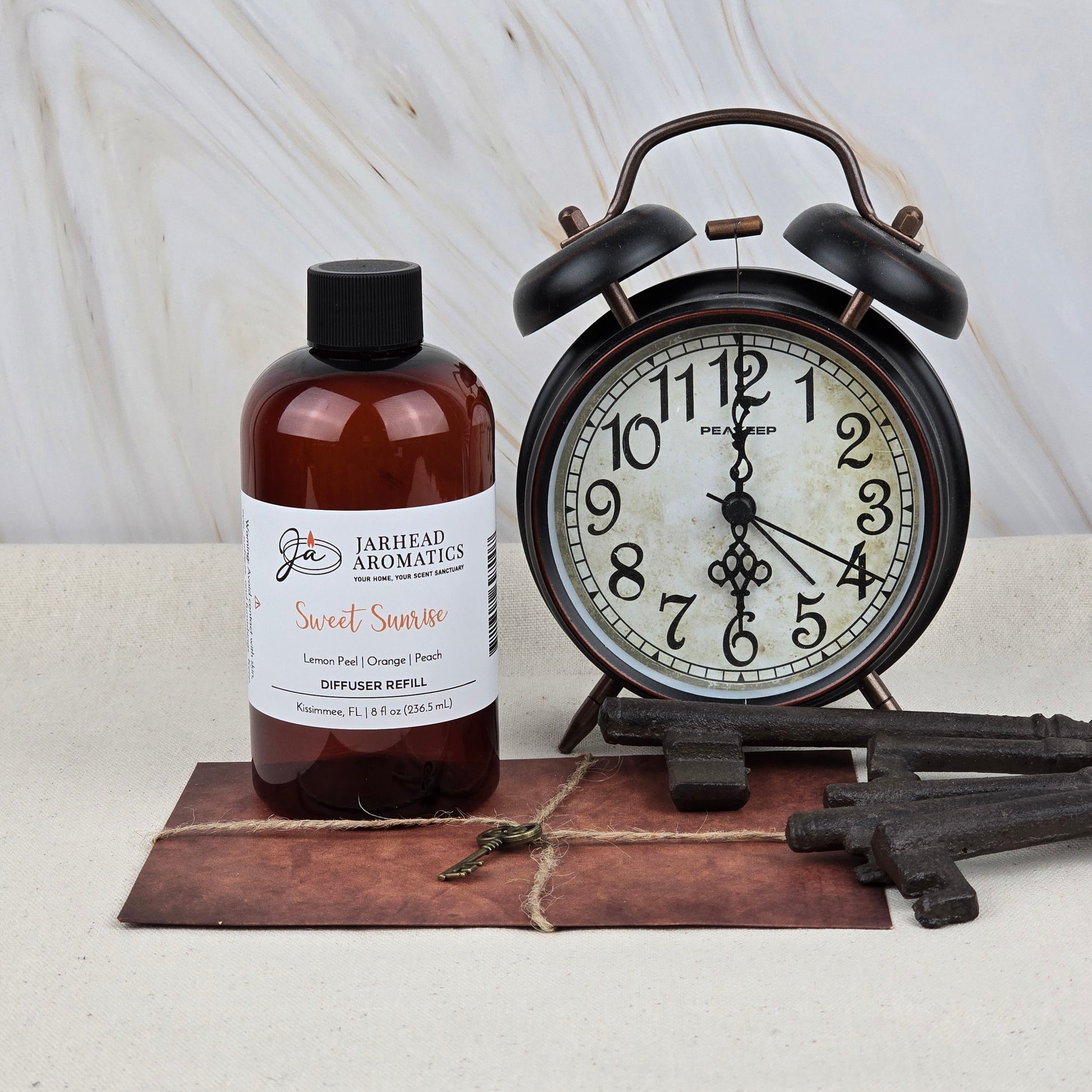 A brown alarm clock sits next to a bottle of Jarhead Aromatics Sweet Sunrise Diffuser Refill | Citrus, Agave & Peach Home Fragrance Oil on a rustic surface with vintage keys.