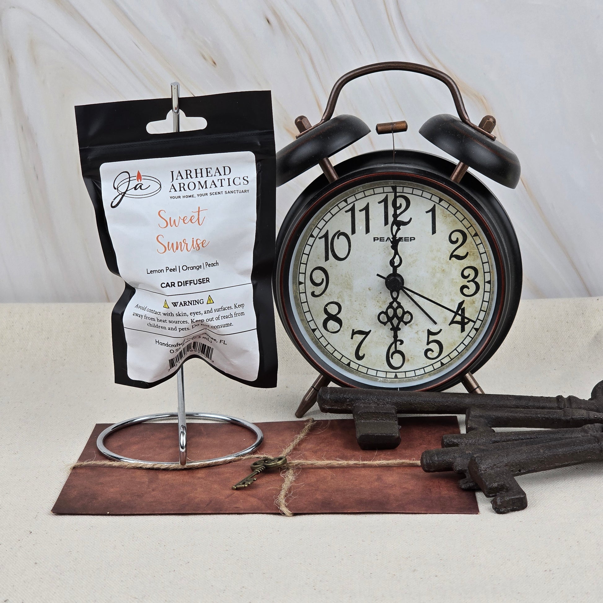 A Sweet Sunrise Car Diffuser | Bright Citrus & Peach by Jarhead Aromatics hangs on a stand beside a vintage black alarm clock at 7:00, with antique keys and a leather item—an elegant, long-lasting car air freshener accent.