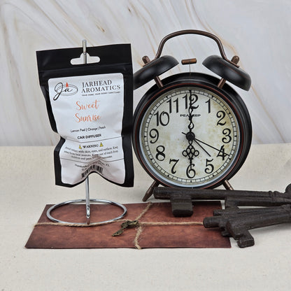 A Sweet Sunrise Car Diffuser | Bright Citrus & Peach by Jarhead Aromatics hangs on a stand beside a vintage black alarm clock at 7:00, with antique keys and a leather item—an elegant, long-lasting car air freshener accent.