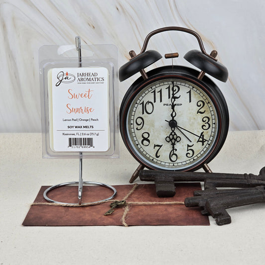 A package of Jarhead Aromatics Sweet Sunrise Soy Wax Melt | Bright Citrus & Peach Wax Melts sits on a metal holder next to a vintage black alarm clock and large keys on a brown envelope atop a beige surface.