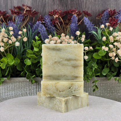 Three rectangular bars of Jarhead Aromatics Tea Tree Handmade Soap - All Natural are stacked on a beige cloth, with lush green leaves and purple and white artificial flowers set against a wooden background.