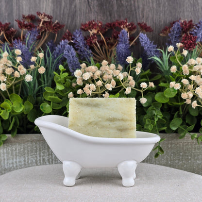 A bar of Jarhead Aromatics Tea Tree Handmade Soap - All Natural rests in a small, white, bathtub-shaped soap dish with artificial flowers and greenery in the background.