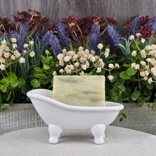 A bar of Jarhead Aromatics Tea Tree Handmade Soap - All Natural rests in a small, white, bathtub-shaped soap dish with artificial flowers and greenery in the background.