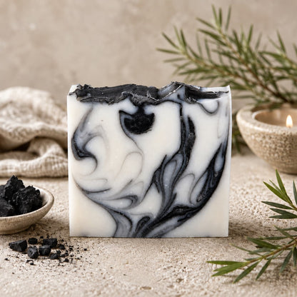 A bar of Jarhead Aromatics Tea Tree + Charcoal Handmade Natural Tallow Artisan Soap, featuring clarifying tea tree and activated charcoal with elegant black and white swirls, rests on a light surface alongside charcoal pieces, a lit candle, a linen cloth, and pine sprigs.