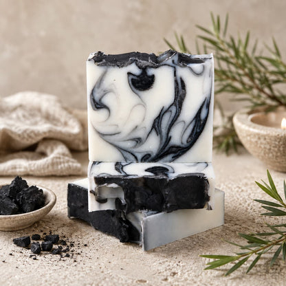 Two bars of Jarhead Aromatics’ Tea Tree + Charcoal Handmade Natural Tallow Artisan Soap, featuring striking black and white swirls, are stacked on a textured surface. Nearby, shards of the clarifying soap, a candle, a sprig of rosemary, and a cloth add to the natural spa-like ambiance.