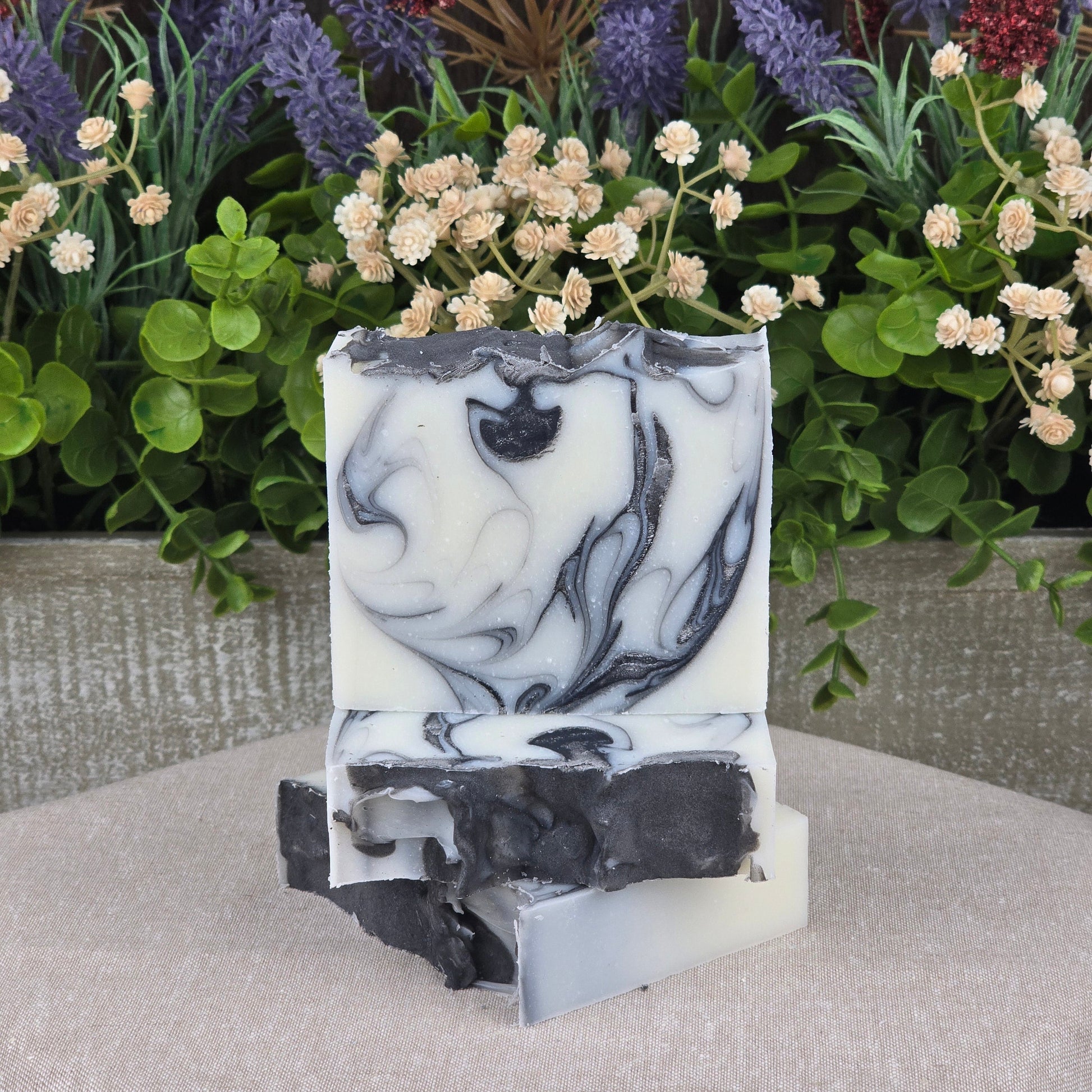 Two Jarhead Aromatics Tea Tree + Charcoal Handmade Natural Tallow Artisan Soap bars with black and white swirls are stacked in front of bright artificial flowers and greenery, creating a soft, natural, rustic vibe.