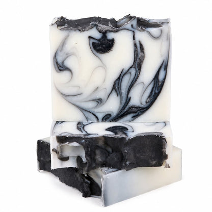 Three bars of Jarhead Aromatics Tea Tree + Charcoal Handmade Natural Tallow Artisan Soap, each showcasing a distinctive marbled black and white swirl, are stacked unevenly against a white background. The clarifying Tea Tree & Activated Charcoal Soap bars feature rustic, uneven edges that emphasize their handcrafted artisan quality.