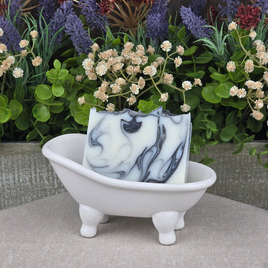 A decorative bar of Jarhead Aromatics’ Tea Tree + Charcoal Handmade Natural Tallow Artisan Soap sits in a small white ceramic bathtub dish, with artificial green leaves and dried purple and red flowers in the background.