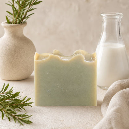 A bar of Jarhead Aromatics Tea Tree Handmade Natural Goat Milk Artisan Soap rests on a light surface, accompanied by a milk bottle, a ceramic vase with greenery, and sprigs of rosemary. The arrangement creates a soft, natural aesthetic.