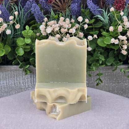 Three bars of Jarhead Aromatics Tea Tree Handmade Natural Goat Milk Artisan Soap, pale green with wavy edges, are stacked before a vibrant display of purple, red, and white artificial flowers on a neutral fabric surface.