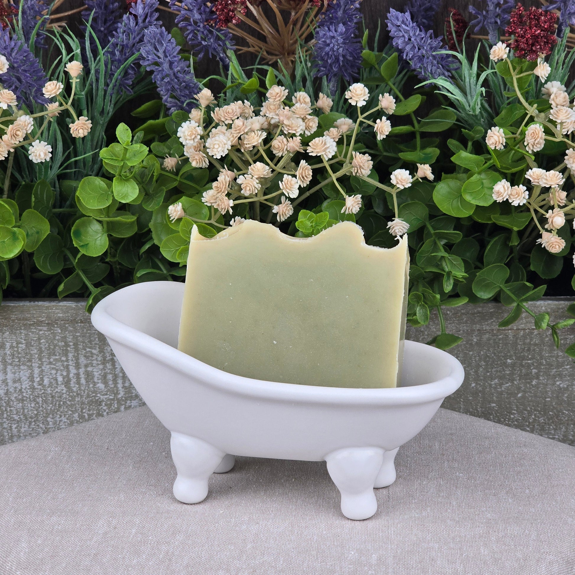 A green bar of Jarhead Aromatics’ Tea Tree Handmade Natural Goat Milk Artisan Soap rests in a white clawfoot bathtub soap dish, with artificial flowers and greenery behind it for a decorative, spa-like display.