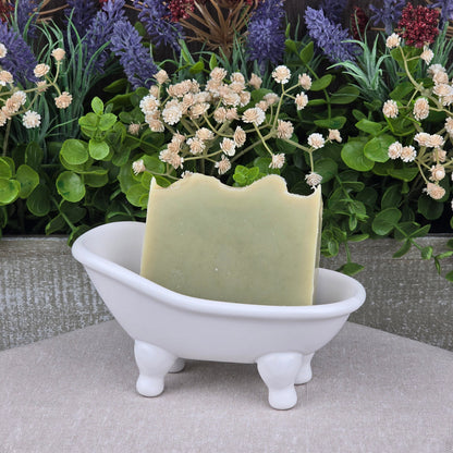 A green bar of Jarhead Aromatics’ Tea Tree Handmade Natural Goat Milk Artisan Soap rests in a white clawfoot bathtub soap dish, with artificial flowers and greenery behind it for a decorative, spa-like display.