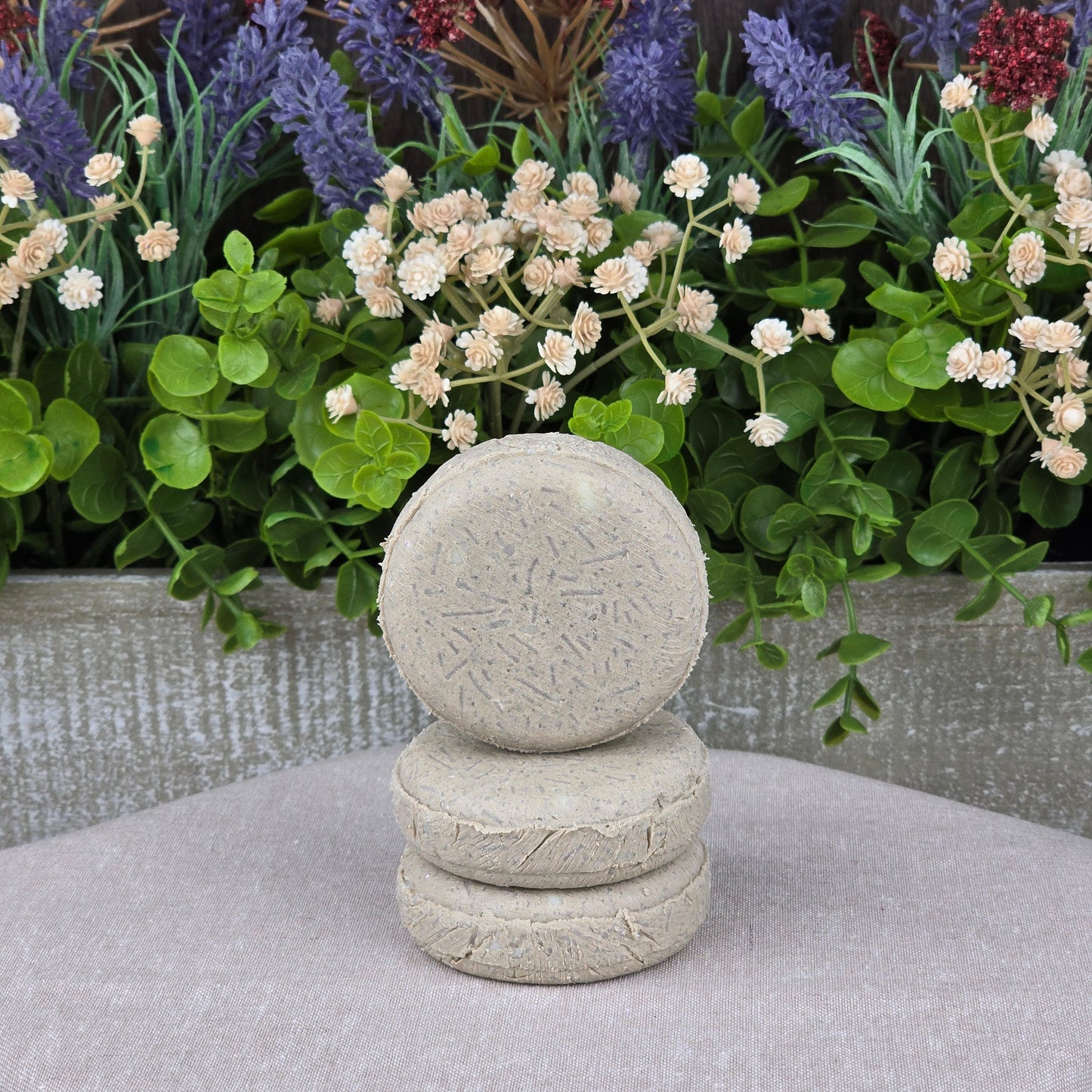 Three beige, textured Tea Tree Handmade Natural Shampoo & Conditioner Bars from Jarhead Aromatics are stacked before green leaves and small white and purple flowers, with a wooden planter and neutral fabric in the background.
